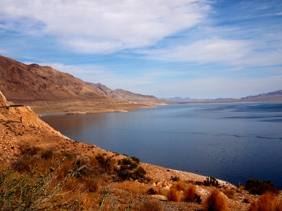 [A large expanse of steel blue lake rimmed by sandstone colored mountains. Plenty of thin white clouds in the blue sky.]